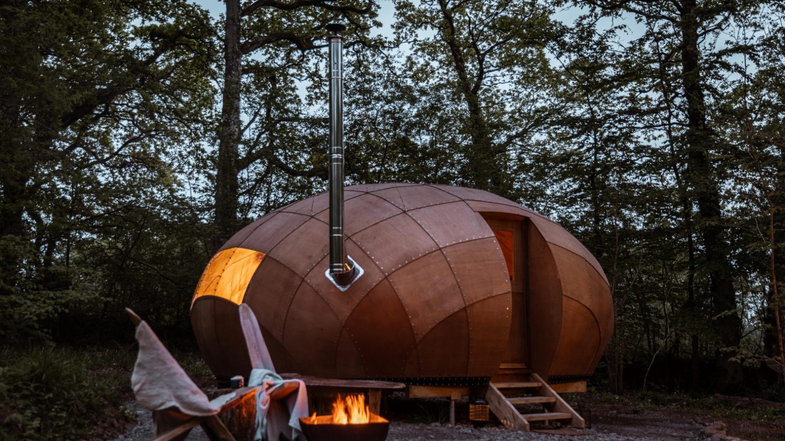 A Honeydown spherical, wooden cabin with a chimney sits in a forest clearing. There's a small staircase leading to its entrance. A glowing fire pit with a wooden chair and draped blanket are in the foreground, surrounded by tall trees at dusk.