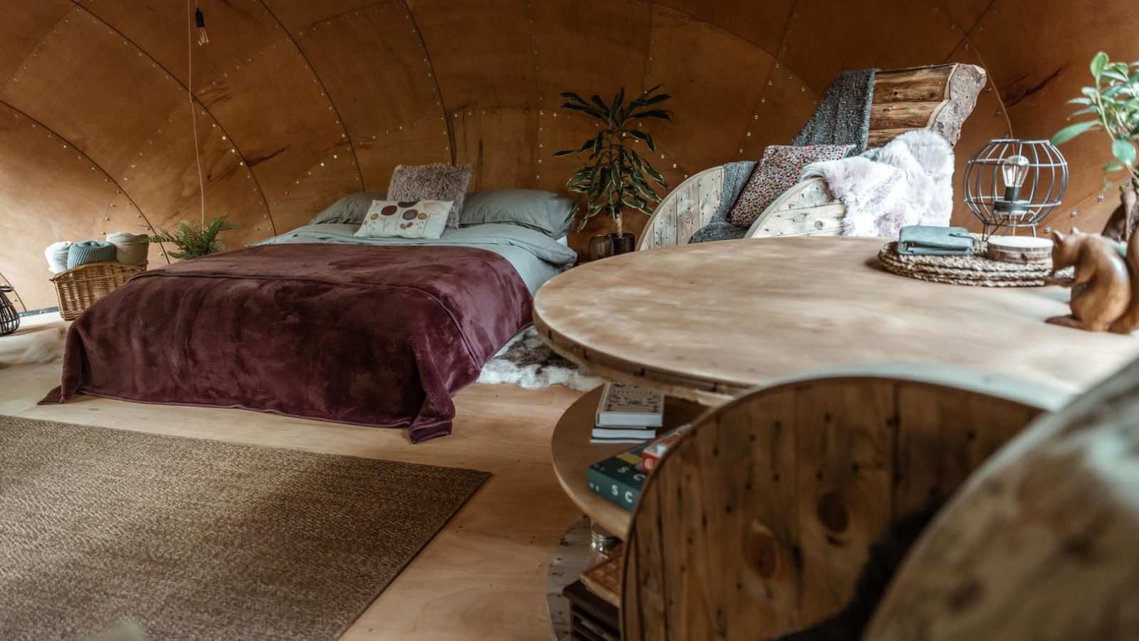 Cozy Honeydown bedroom with a curved wooden ceiling, featuring a bed with a maroon blanket, a woven rug, a plant, a round wooden table with books on a lower shelf, and cushioned seating. The space exudes warmth and rustic charm.