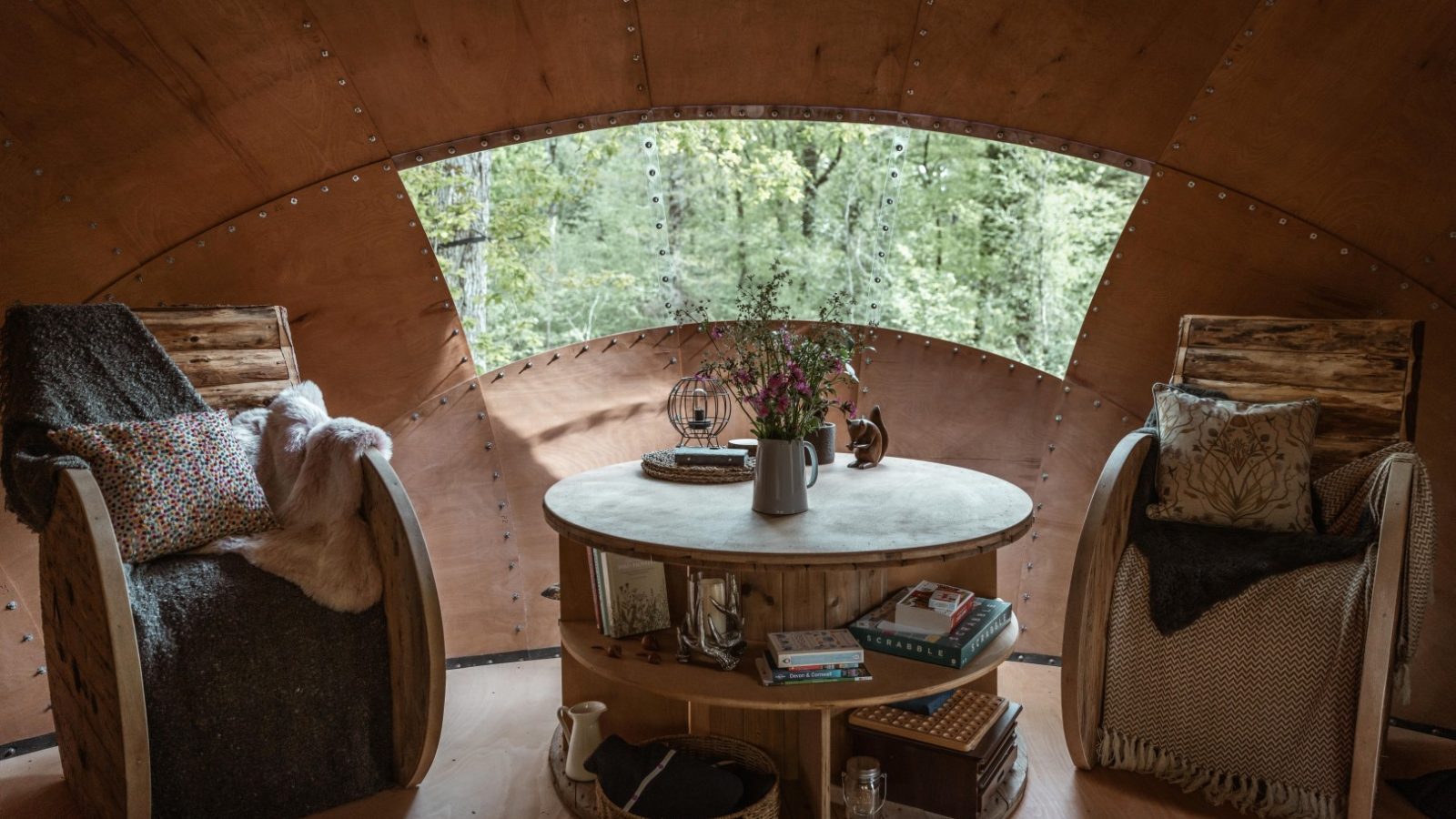 Cozy Honeydown room with wooden chairs, a round table adorned with books and flowers, and a large window overlooking a stunning forest landscape.