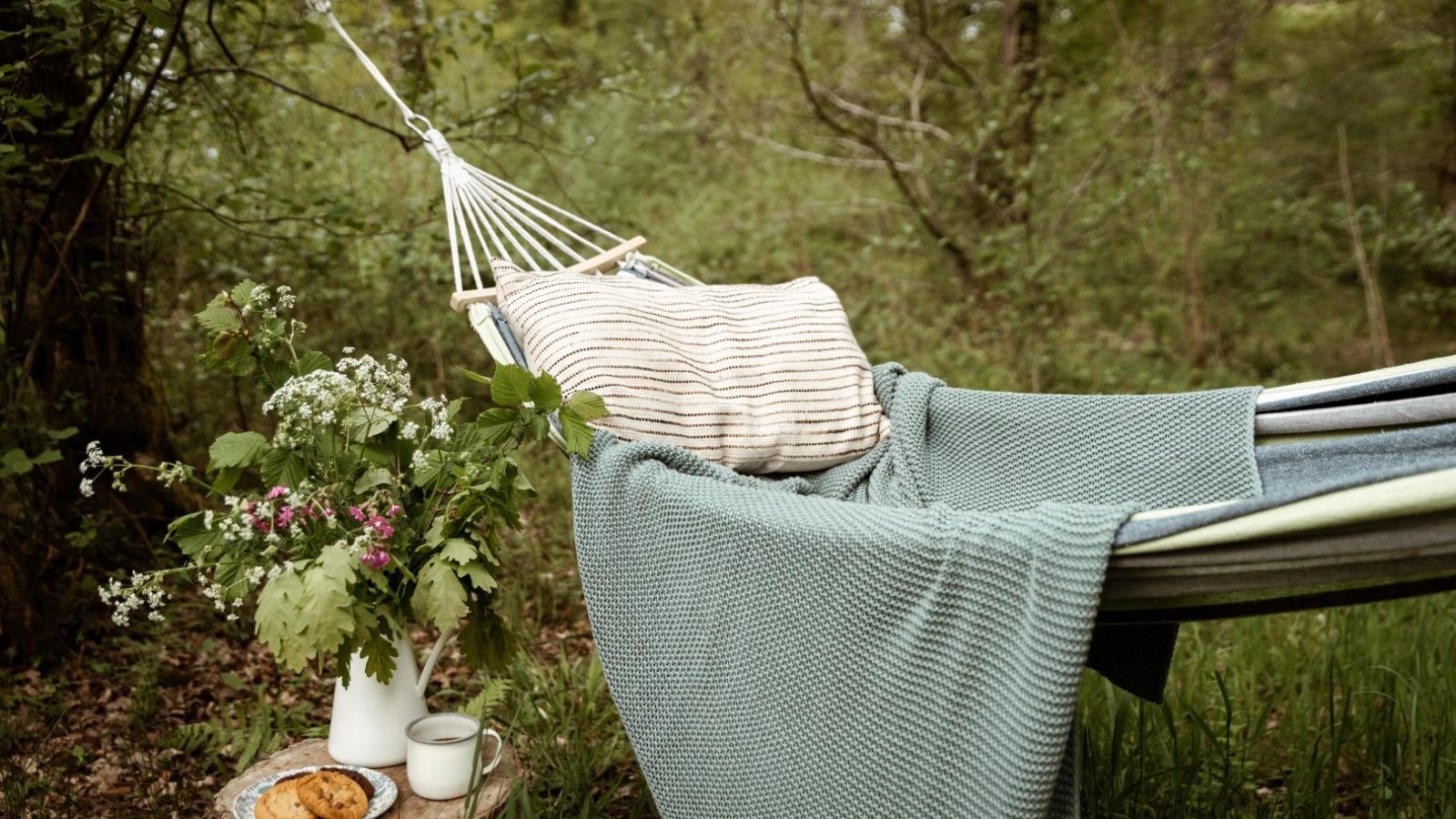 A cozy Honeydown hammock hangs in a lush forest, draped with a green blanket and striped pillow. Nearby, a tree stump serves as a table, holding a white vase of wildflowers, a mug, and a plate with cookies. The scene exudes a peaceful, relaxing atmosphere.