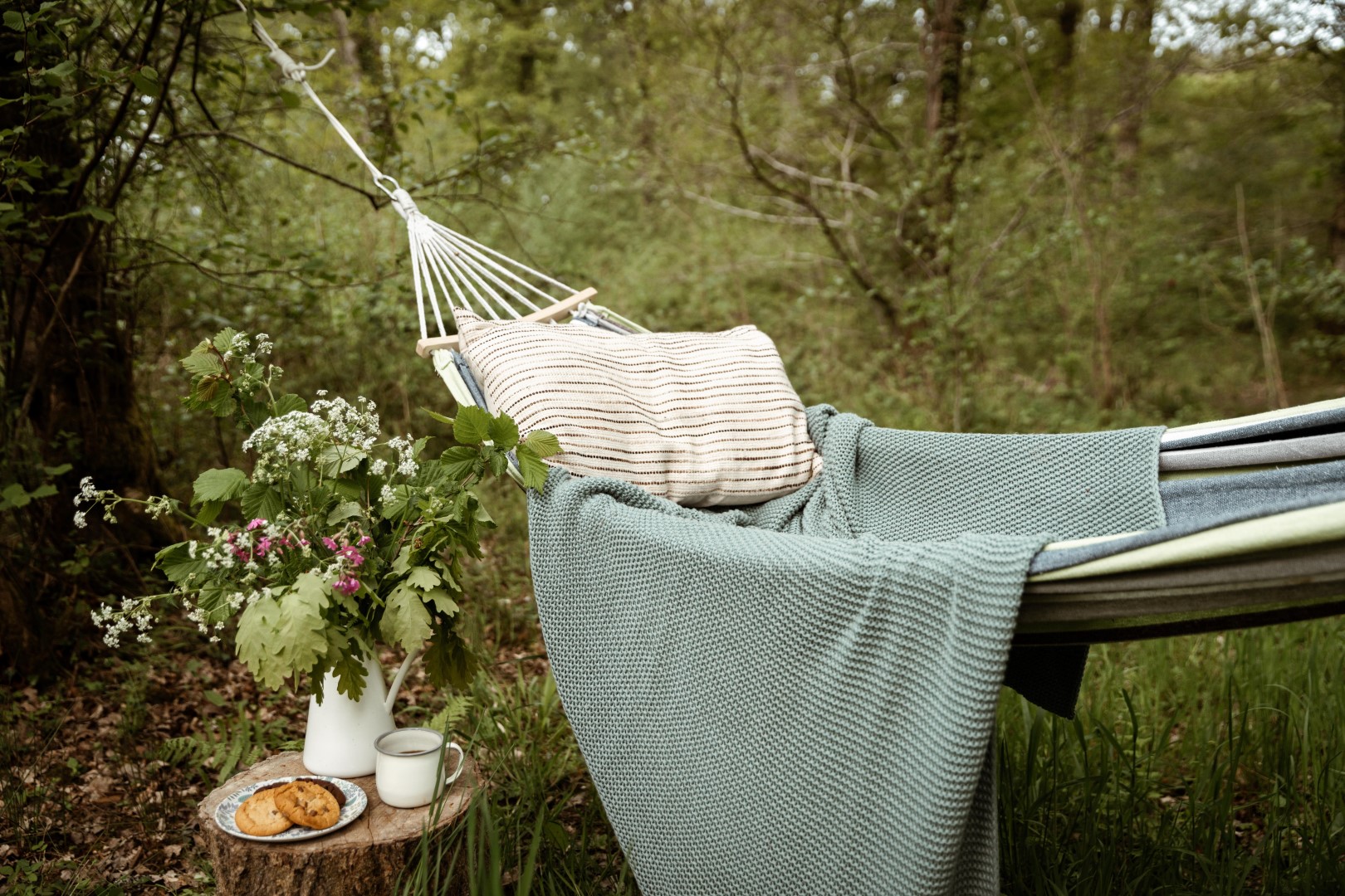 A hammock is tied between trees in a forest setting, adorned with a striped pillow and a blue blanket. Next to it, a tree stump holds a white pitcher of flowers, a white mug, and a plate with cookies. Perfect for your holiday let promotion on social media to increase engagement, the backdrop brims with lush green foliage.