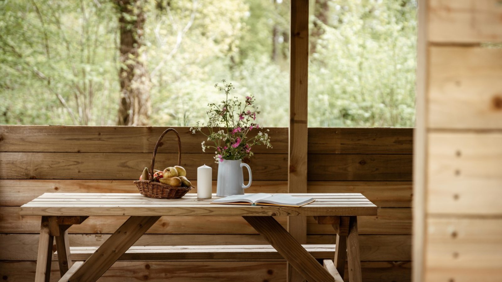 A wooden picnic table sits under a covered outdoor shelter in Honeydown. On the table, there is a basket of fruit, two candles, a vase with wildflowers, and an open book. The background features lush green forest scenery.