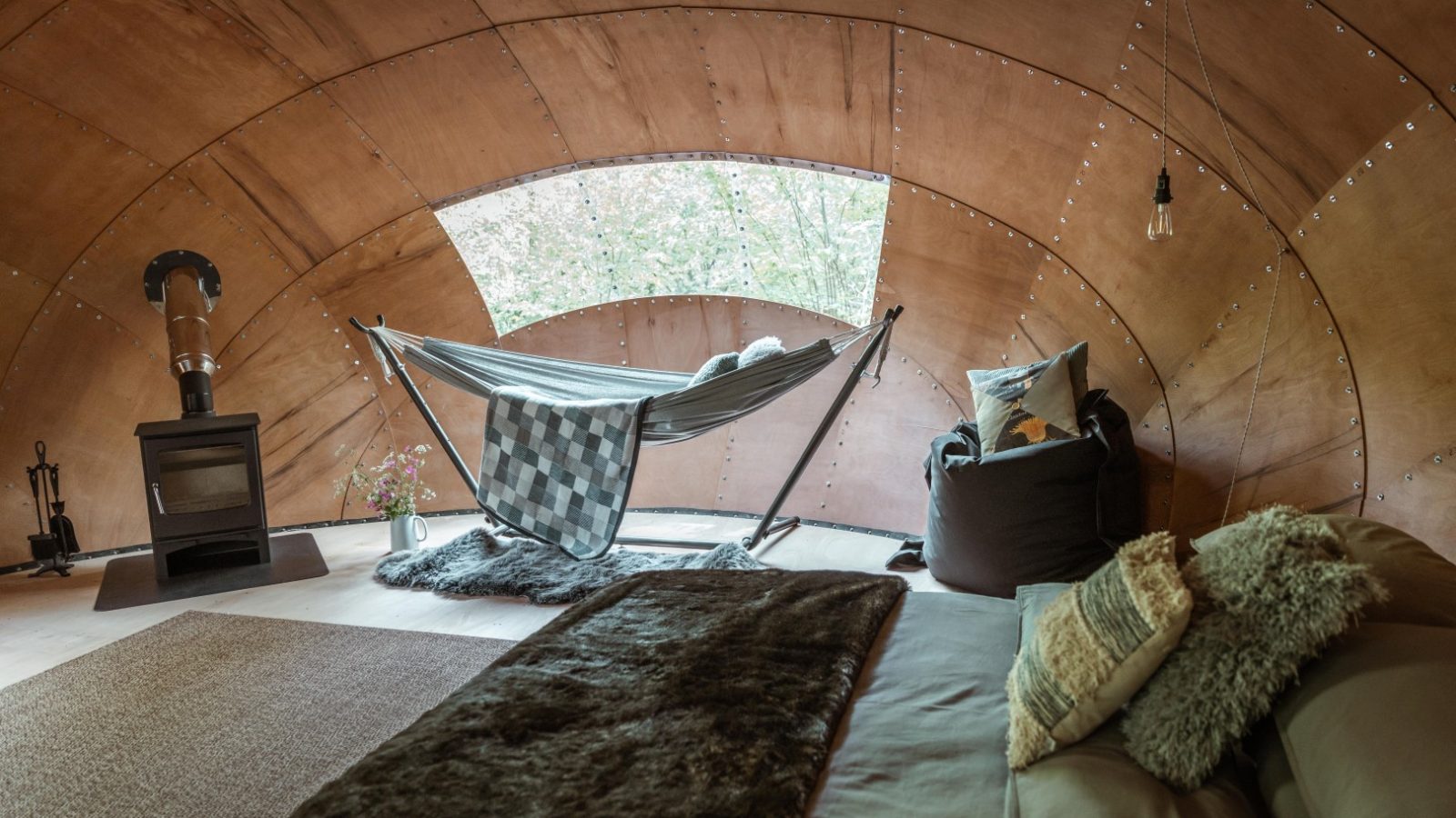 A cozy interior of a Honeydown wooden cabin with a large window. The room features a hammock with a black and white checkered blanket, a bean bag chair, and a bed with pillows and a fur throw. A wood-burning stove sits in the corner with a kettle on top.