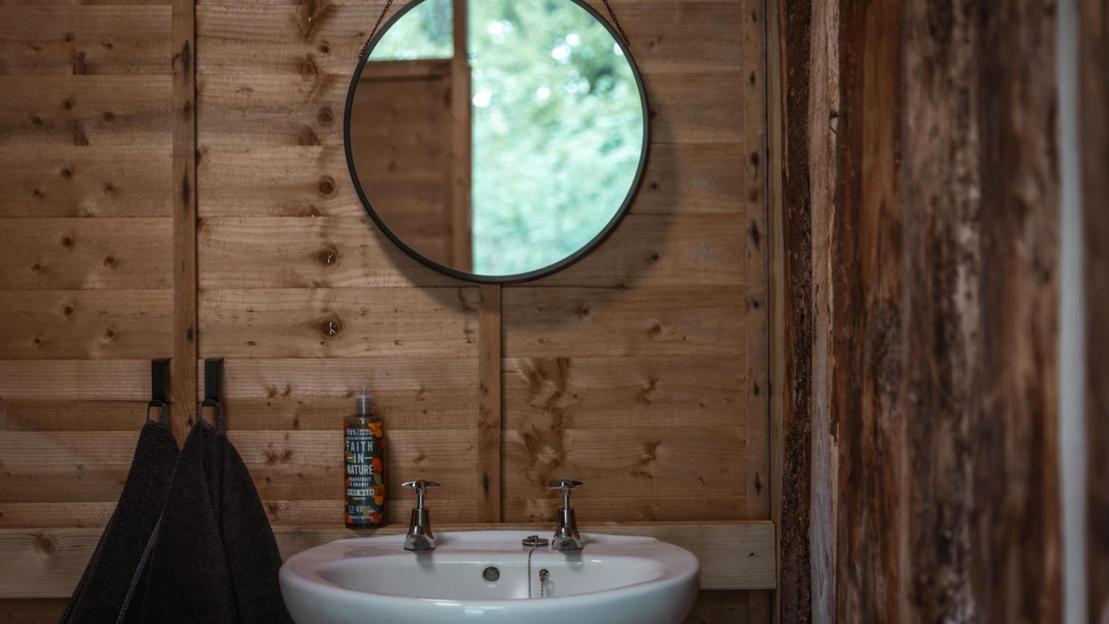 A rustic bathroom in Honeydown features a wooden wall, a round mirror hanging above a white basin with chrome faucets, and two dark towels on hooks beside the sink. A can of shaving cream is placed on the ledge behind the sink. A window reflects greenery in the mirror.