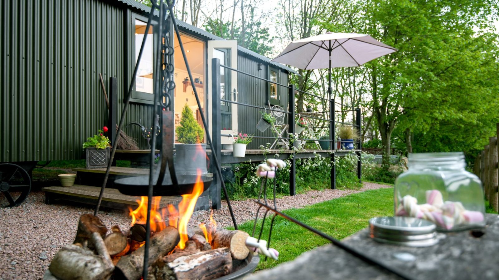 A cosy outdoor scene features a fire pit with burning logs and marshmallows on skewers beside a rustic green cabin, evoking the charm of rural retreats. A patio with a parasol, potted plants, and chairs is visible, surrounded by lush greenery and trees—hop pickers retreat