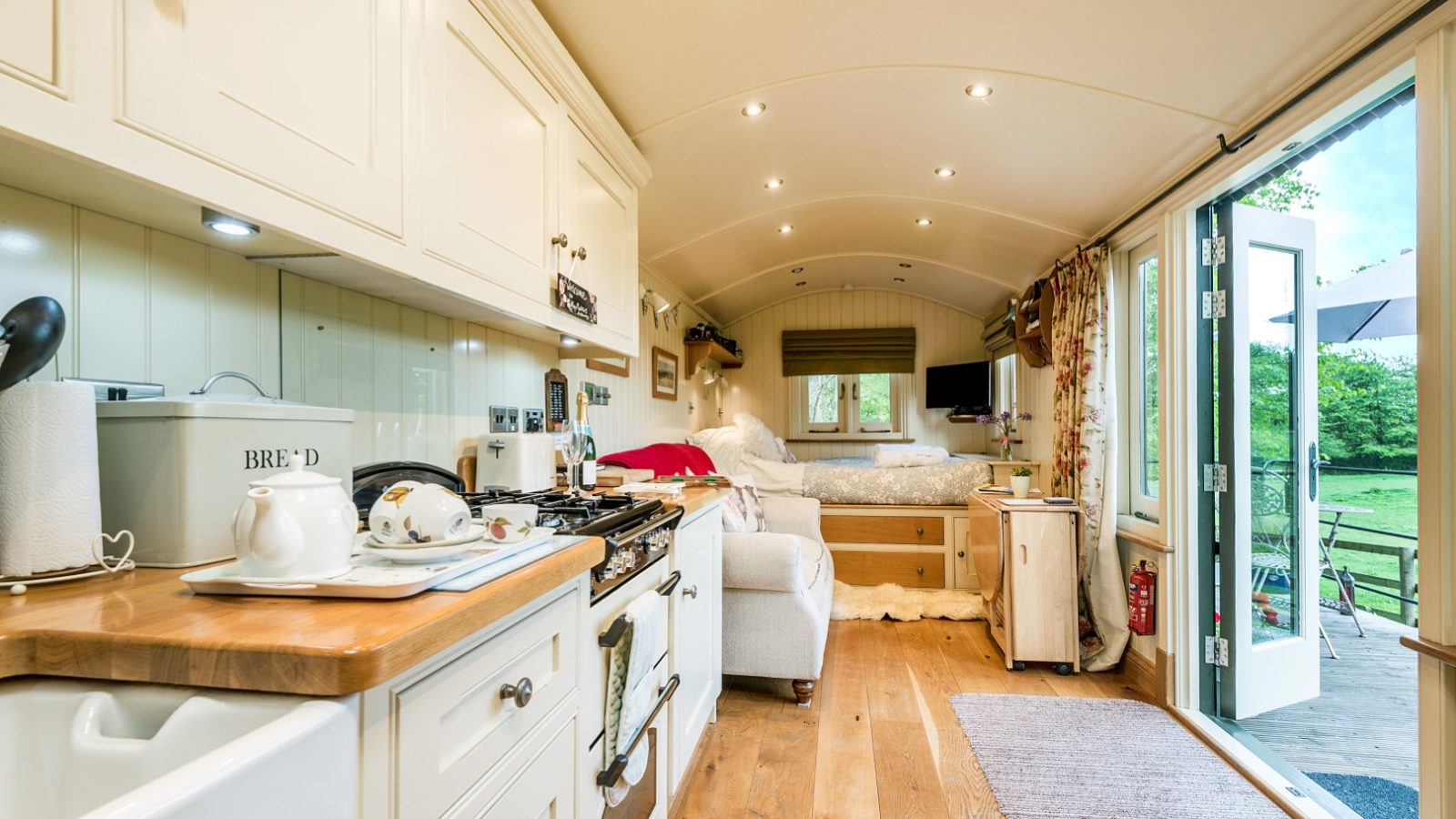 Cozy tiny home interior with wooden floors, white cabinets, and a small kitchenette on the left. A plush sofa sits in the center, leading to a bed in the back. Open doors reveal a grassy outdoor area with a table and chairs, perfect for hop pickers seeking rural retreats.