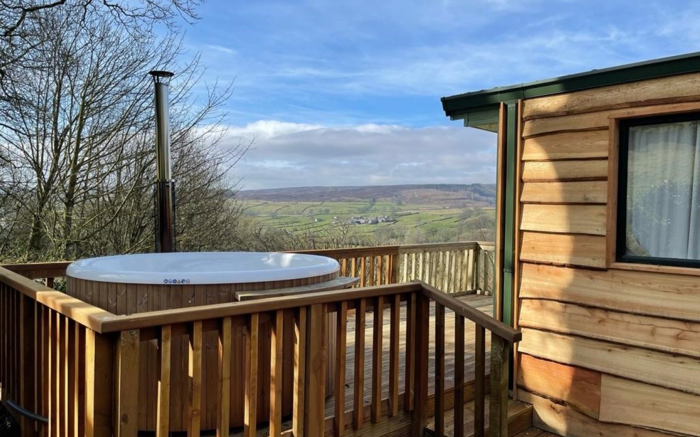 A wooden cabin deck with a hot tub at Howgill Lodge overlooks a scenic countryside landscape. The view features rolling hills, fields, and a partly cloudy sky. Bare trees frame the deck, adding a natural touch to the rustic outdoor setting.