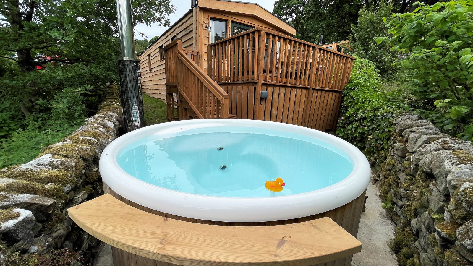 An inviting outdoor hot tub with clear water and a rubber duck floating in it sits between a stone wall and a wooden deck, which leads up to the cozy Howgill Lodge cabin surrounded by trees and greenery.