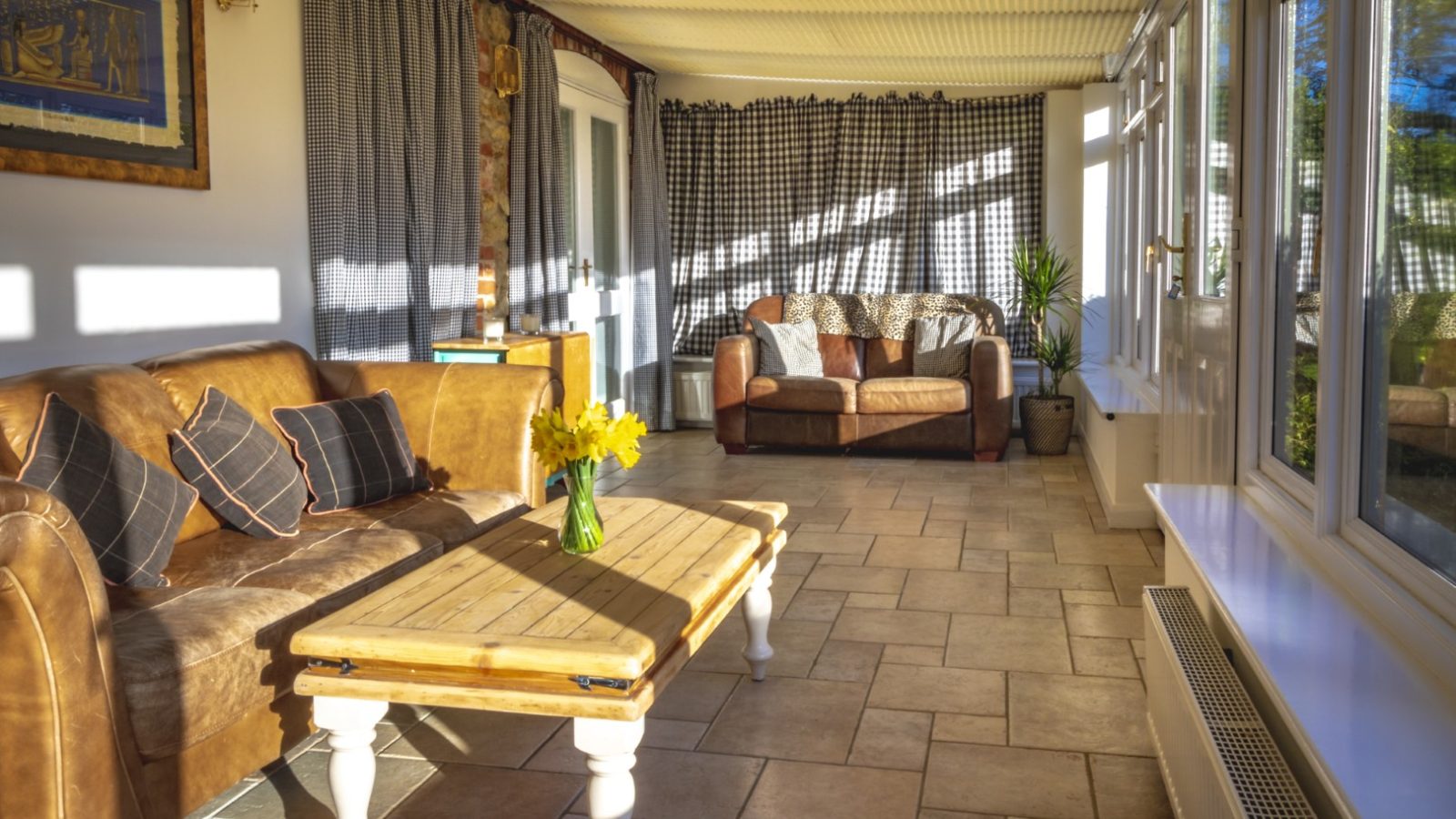 A bright sunroom at Pennard Farm Cottages features tiled flooring, a brown leather sofa adorned with blue and beige cushions, and a wooden coffee table with a vase of yellow flowers. A second sofa sits near the back, against checkered curtains. Large windows let in ample sunlight.