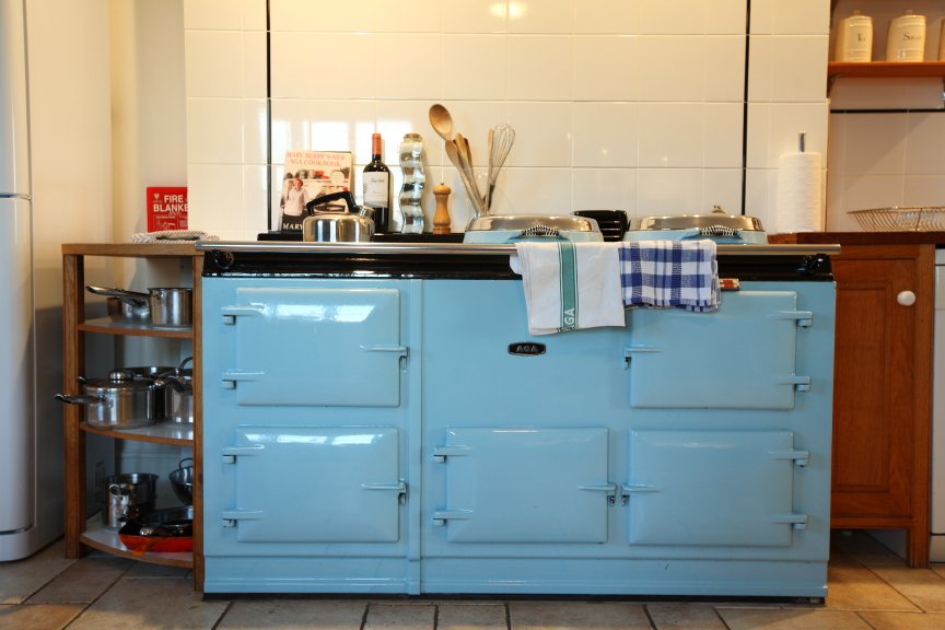 A light blue vintage stove with multiple doors is centered in a kitchen at Pennard Farm Cottages. Various cooking utensils and bottles are on a shelf behind the stove. A white tile backsplash contrasts with the stove, and a silver drying rack with dishes is visible on the right.