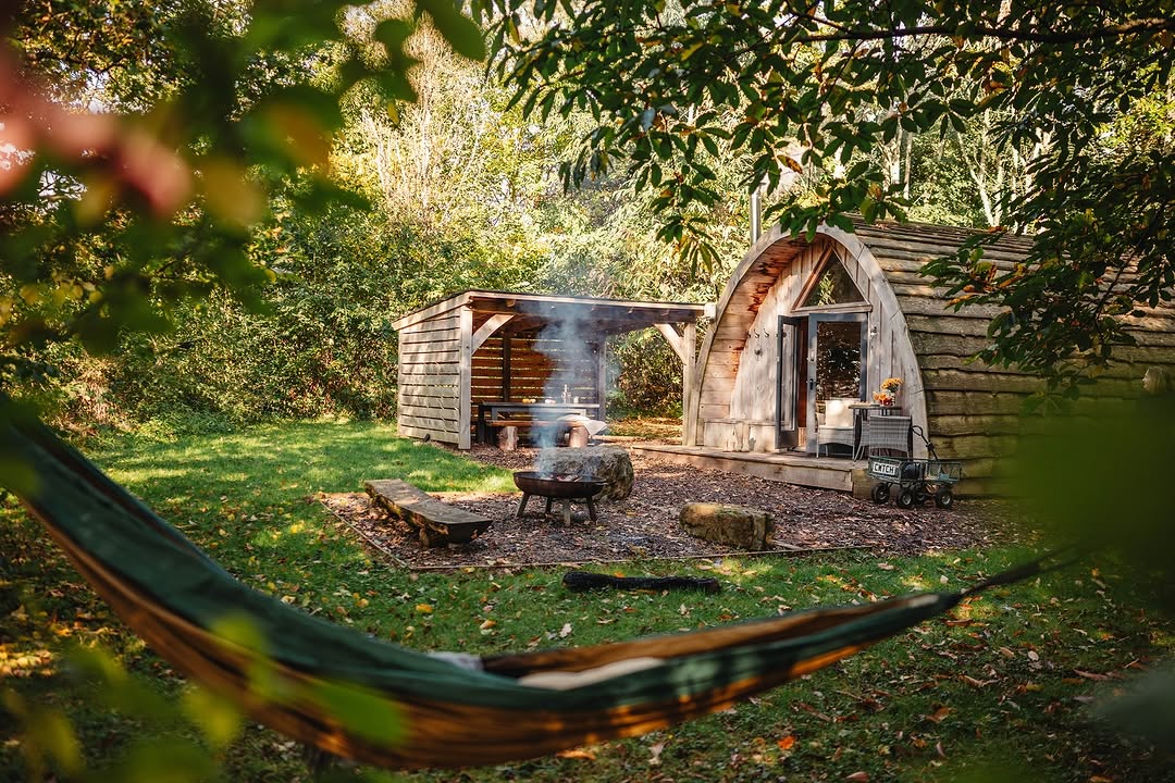 Woodland cabin with an arched roof in Ffynnonau, featuring a hammock, fire pit, and sheltered seating area outside.