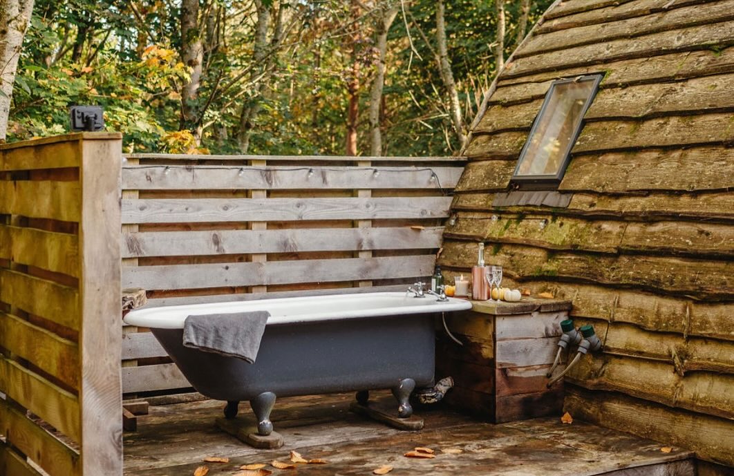 Outdoor clawfoot bathtub on wooden deck of Ffynnonau's Woodland Cabins, surrounded by rustic fence and autumn foliage.