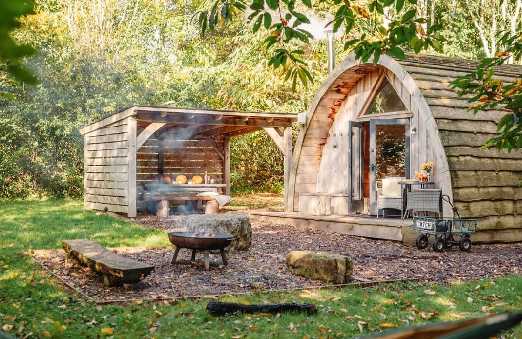 A woodland cabin with a curved roof sits in the forest. A fire pit warms a picnic area with pumpkins and a small trolley nearby.