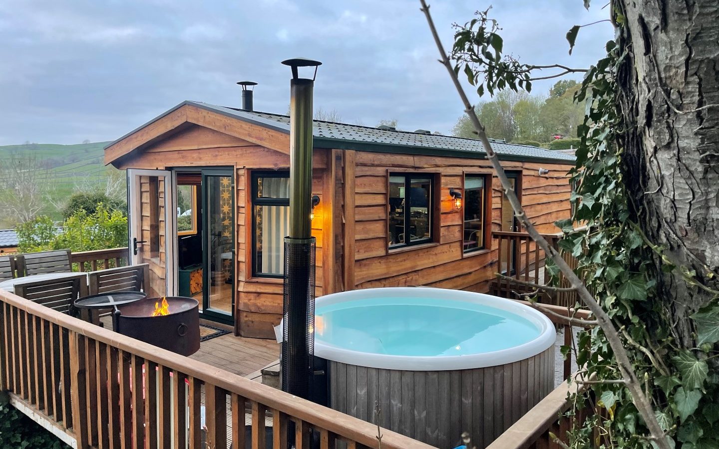 Nestled among trees with rolling hills under a cloudy sky, this charming lodge features a wraparound deck with a hot tub, tall outdoor heater, and small fire pit. Experience serene accommodation at Howgill Lodge in your cozy wooden cabin haven.