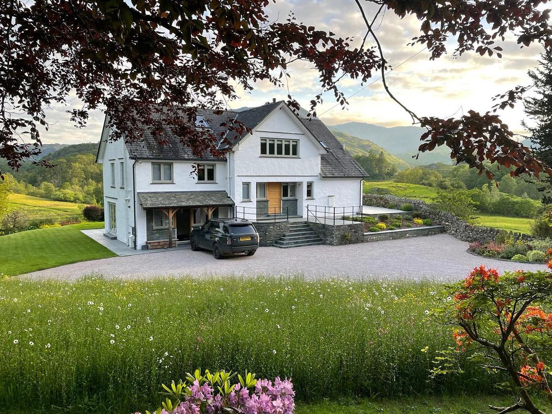 A picturesque white two-story house, known as Lowfield House, has a black car parked in front. It's surrounded by lush greenery and wildflowers. Trees frame the scene, while a serene landscape with hills unfolds under a cloudy sky.