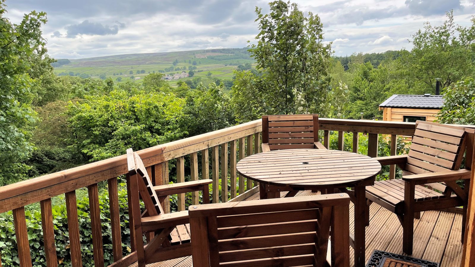 A wooden balcony with a round table and four chairs overlooks the lush green valley of Howgill Lodge, with rolling hills in the background. The sky is cloudy, and the scene is surrounded by dense foliage, creating a serene and scenic view.