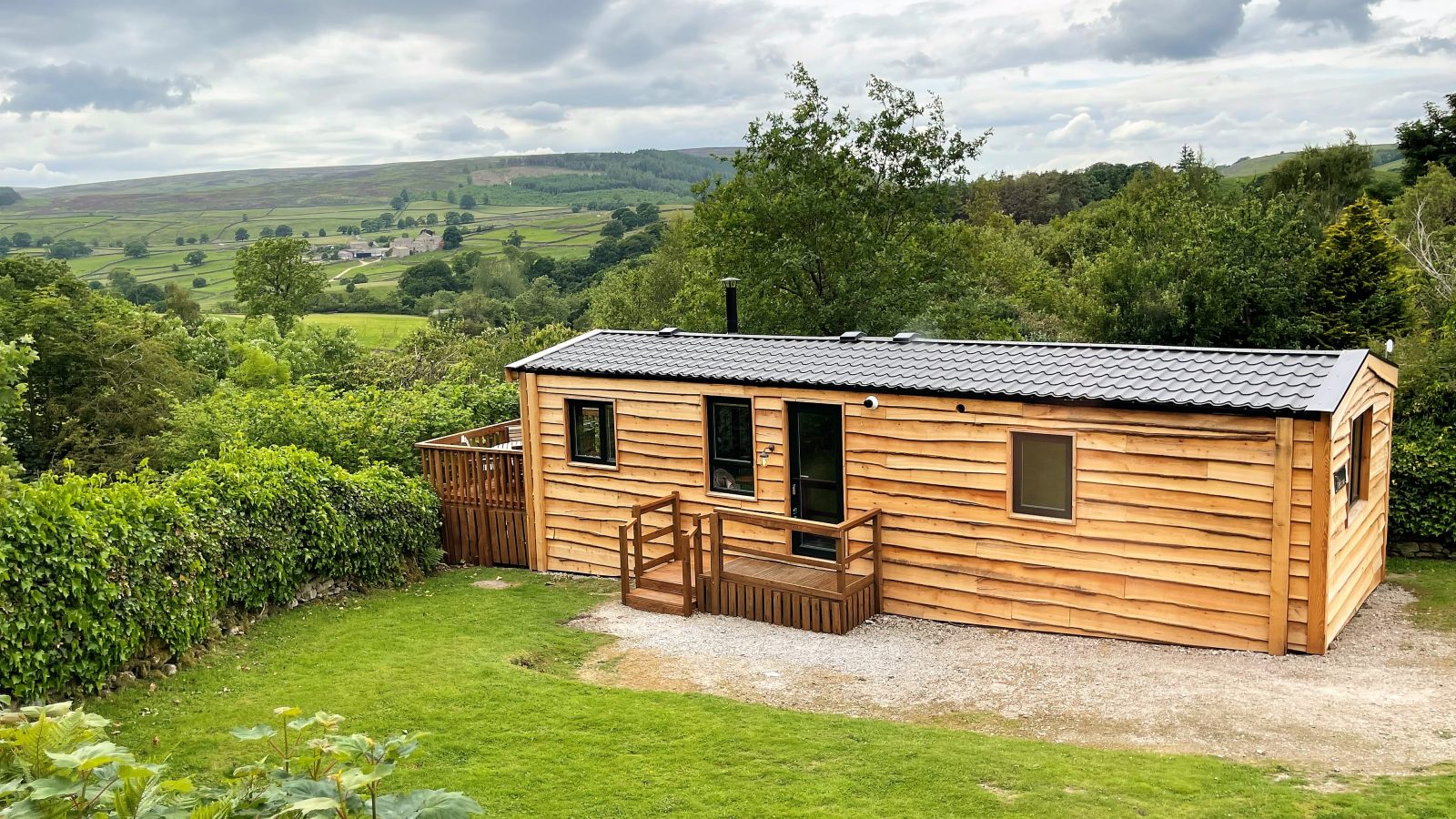 A small wooden cabin with a deck, surrounded by lush greenery and overlooking a scenic landscape of rolling hills and valleys at Howgill Lodge. The sky is partly cloudy, and the cabin is set on a gravel area with a manicured lawn in the foreground.