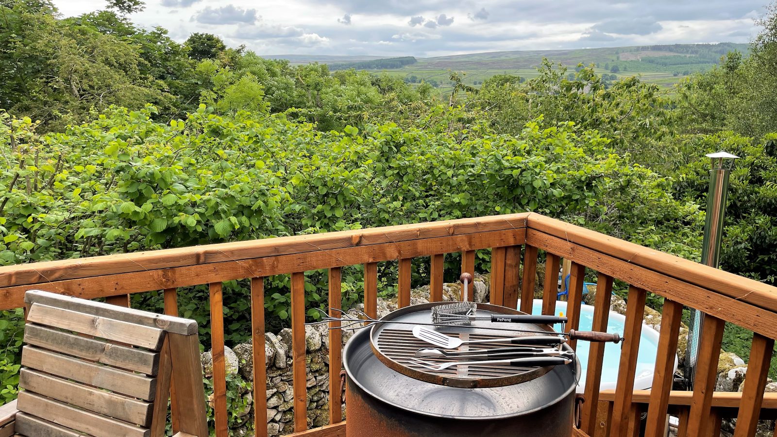 A scenic outdoor deck at Howgill Lodge with a wooden railing overlooks a lush green landscape. The deck features a barbecue grill with cooking utensils on top and two wooden chairs. The sky is cloudy, and the view includes grassy hills and dense trees.