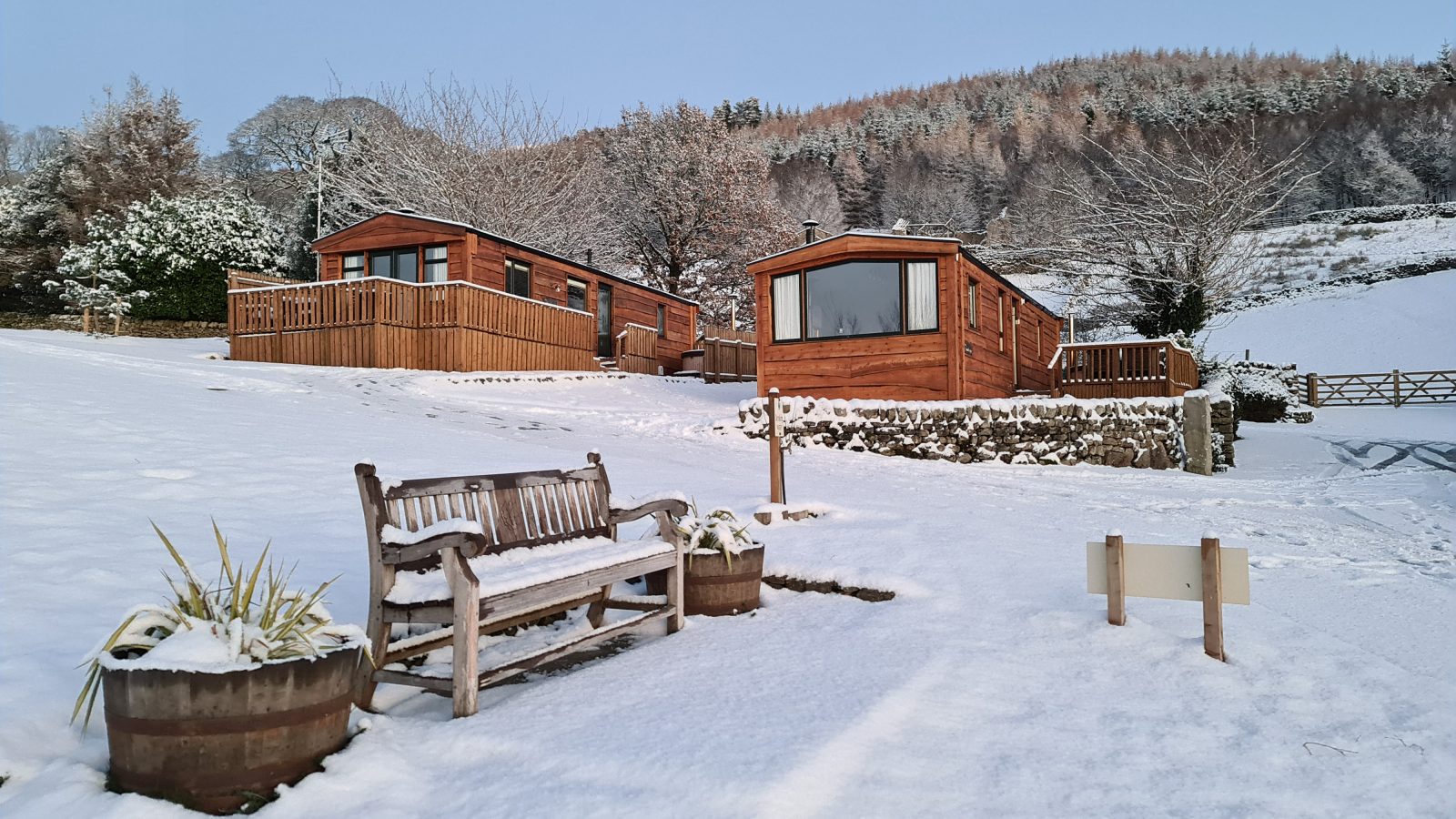 A serene, snow-covered landscape at Howgill Lodge features two wooden cabins on a hillside. A wooden bench and planters sit in the foreground, blanketed in snow. Trees and hills in the background are also dusted with snow, creating a peaceful winter scene.