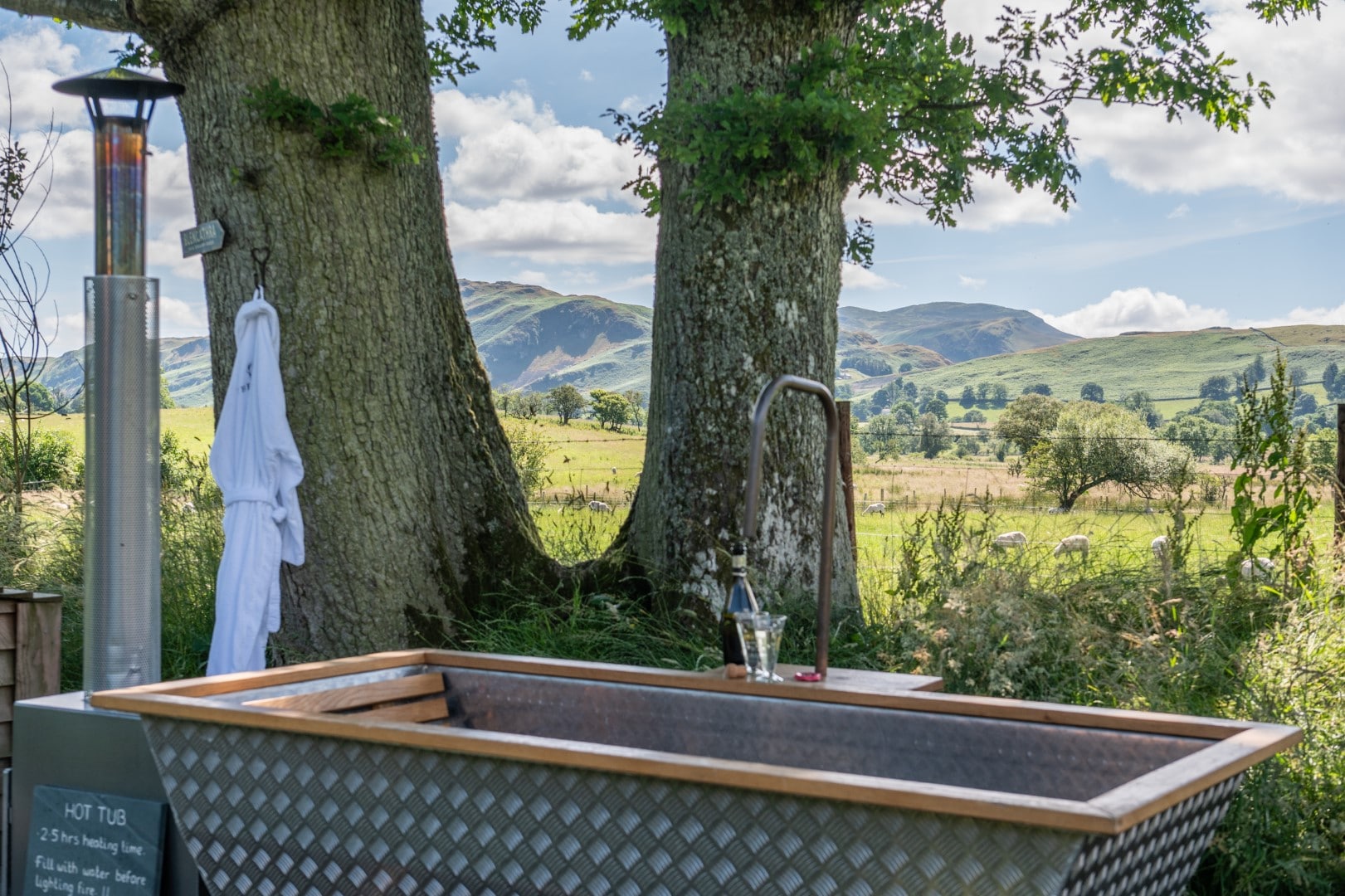 An outdoor hot tub with a metallic finish is set between two large trees, overlooking a scenic landscape of green fields and distant hills. A white robe hangs from a hook on a tree, and a bottle of wine with glasses is placed on the rim of the tub.