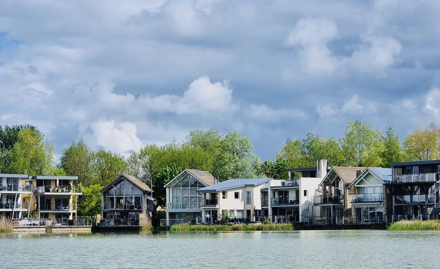 A row of modern houses with large windows and balconies line a peaceful lakeshore, illuminated softly by lamplight. The houses are surrounded by greenery, and the lake reflects the calm sky filled with fluffy clouds.