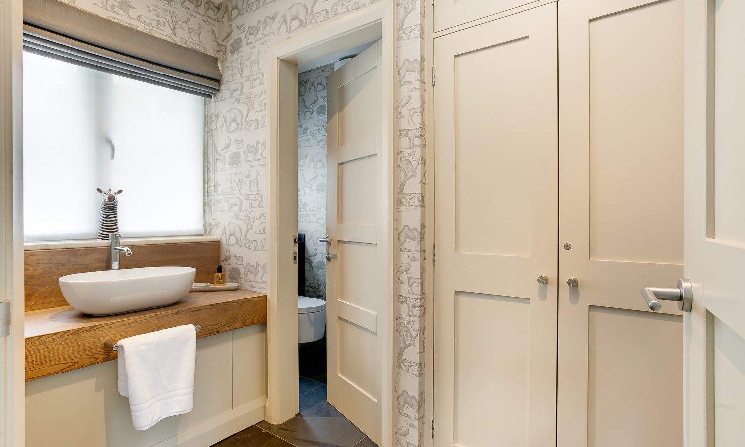 In a small Lowfield House bathroom, a modern white sink rests on a wooden counter. Above, a frosted window holds a zebra figurine. The walls showcase animal-patterned wallpaper, with a cabinet on the right and a door leading to the toilet.