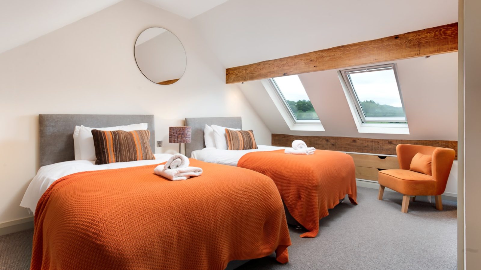 The cozy Lowfield House bedroom features two single beds covered in orange blankets, topped with neatly folded towels. A round mirror adorns the wall, while skylight windows flood the space with natural light. An inviting orange armchair sits beside the window.