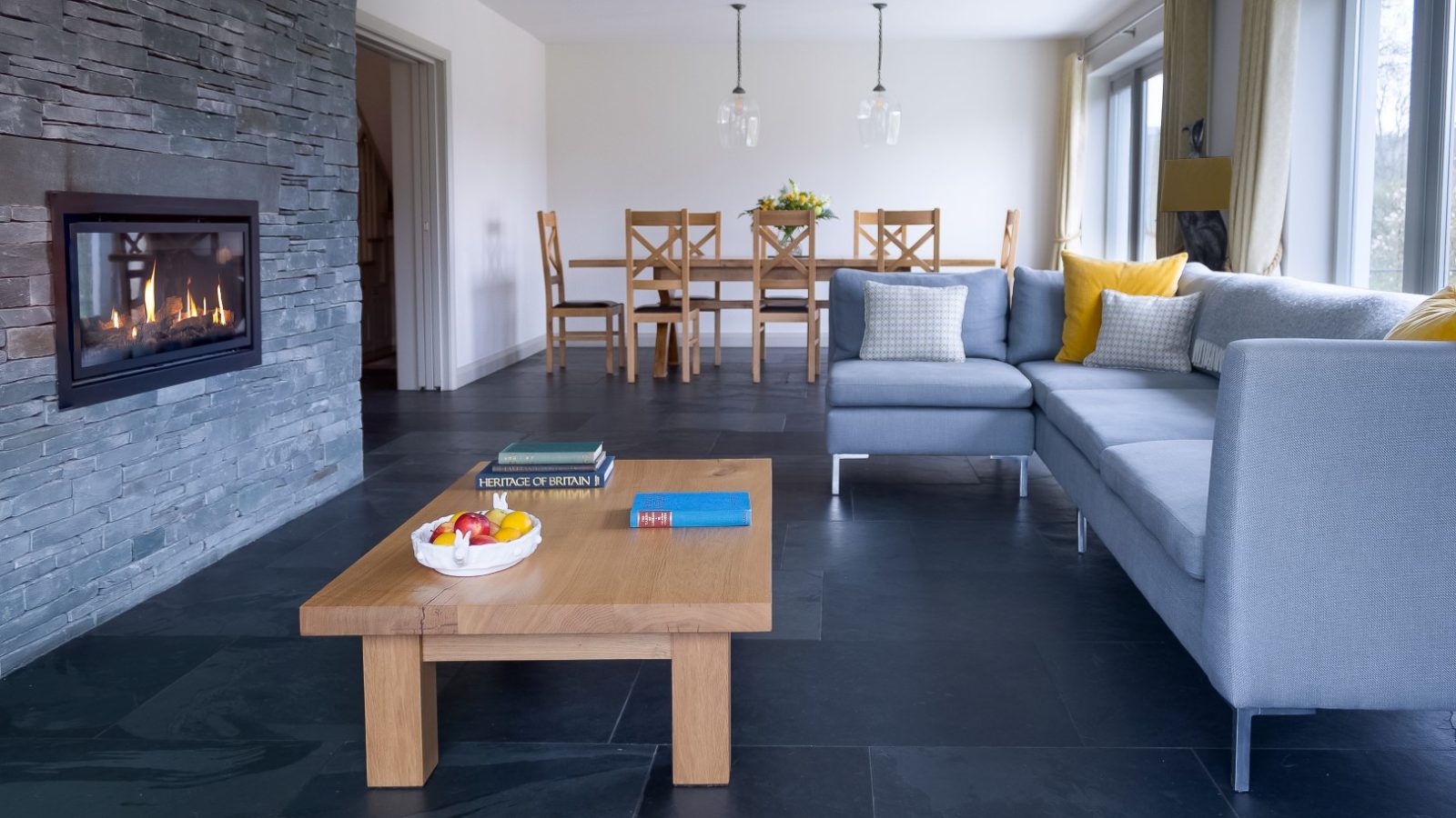 In the heart of Lowfield House, a modern living room features a gray sectional sofa adorned with yellow and white pillows. A wooden coffee table displays books and fruit, while a stone fireplace adds warmth. The dining area with wooden chairs extends invitingly in the background.