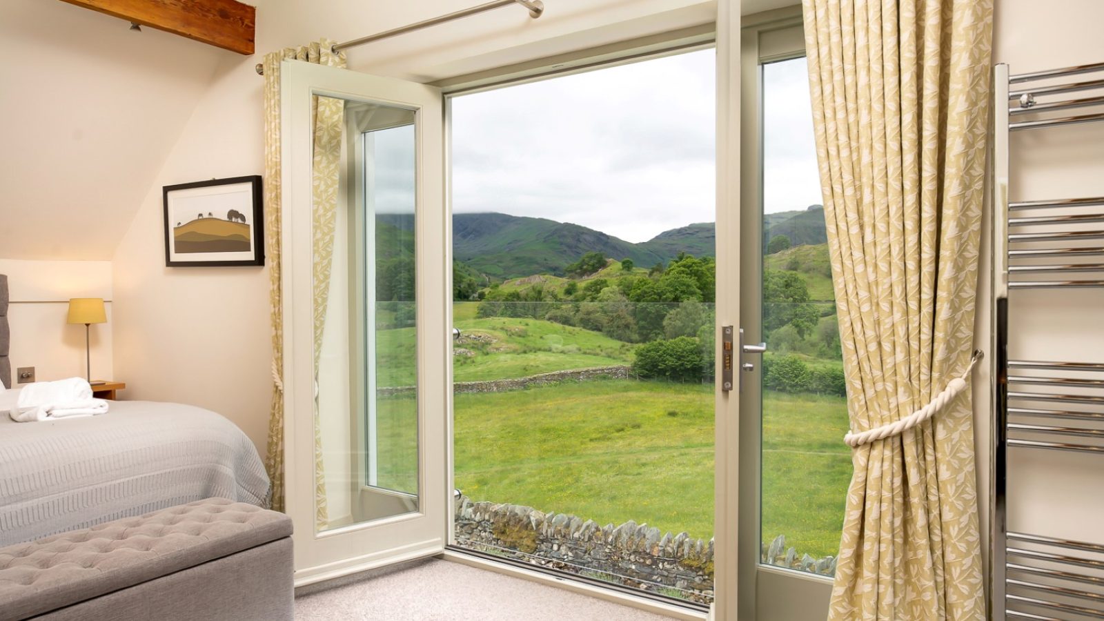 A cozy bedroom in Lowfield House features a large open door that leads to a balcony overlooking a scenic landscape with rolling green hills and a stone fence. The room includes a neatly made bed with a wooden beam ceiling accent, soft lighting, and beige curtains.