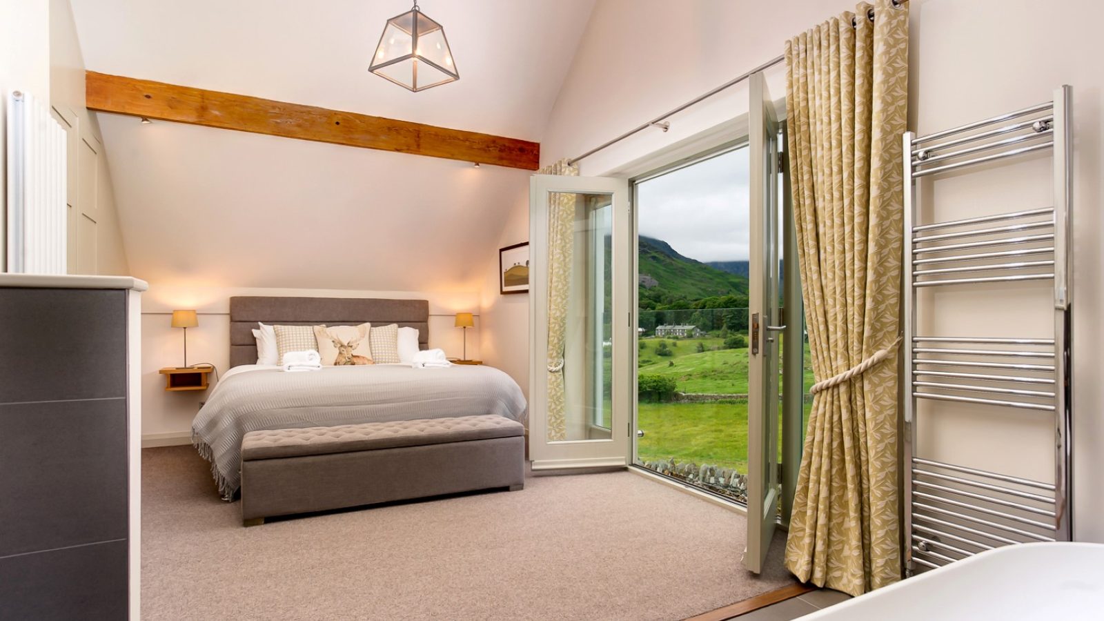 At Lowfield House, a spacious bedroom boasts a large bed with gray linens, a wooden beam ceiling, and a geometric light fixture. Glass doors reveal verdant hills. Gold curtains frame the view while a heated towel rack and glimpses of the bathtub add luxurious touches.