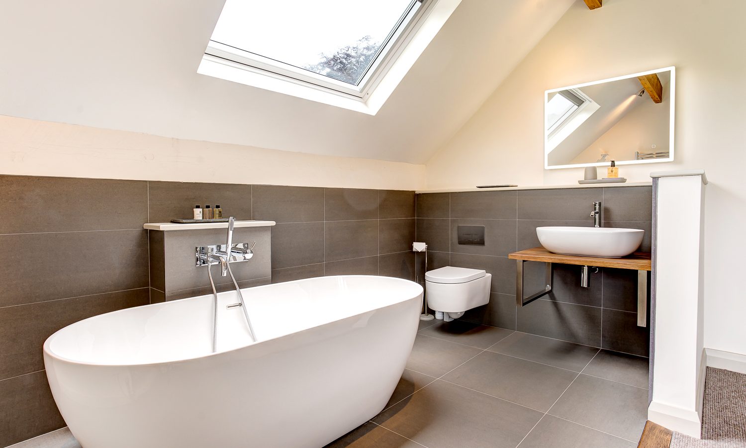 The modern bathroom at Lowfield House boasts a sloped ceiling with a skylight. It features a freestanding white bathtub, wall-mounted sink with a wooden countertop, and square mirror. Neutral tones dominate the design, with gray floor and wall tiles offering serene sophistication.