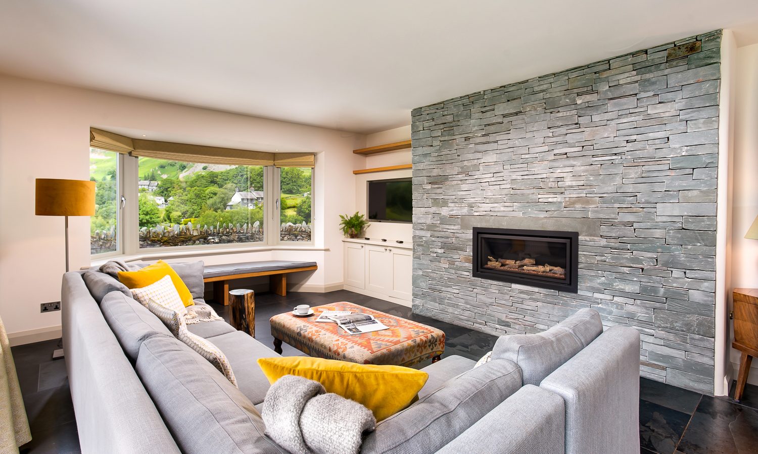 A modern living room in Lowfield House features a large gray sectional sofa adorned with yellow and white pillows, facing a wall with a built-in fireplace and mounted TV. The back wall boasts gray stone cladding, while a bay window offers a scenic view of greenery and hills.