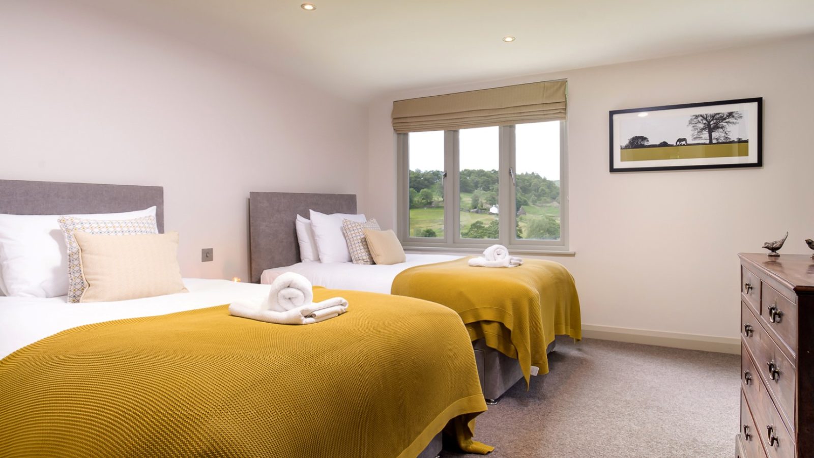 A cozy bedroom at Lowfield House features two neatly made twin beds, each adorned with mustard-colored throws and white pillows. Towels rest on the beds, while a window reveals a scenic view of green fields. A landscape painting hangs above a wooden dresser, adding charm to the space.