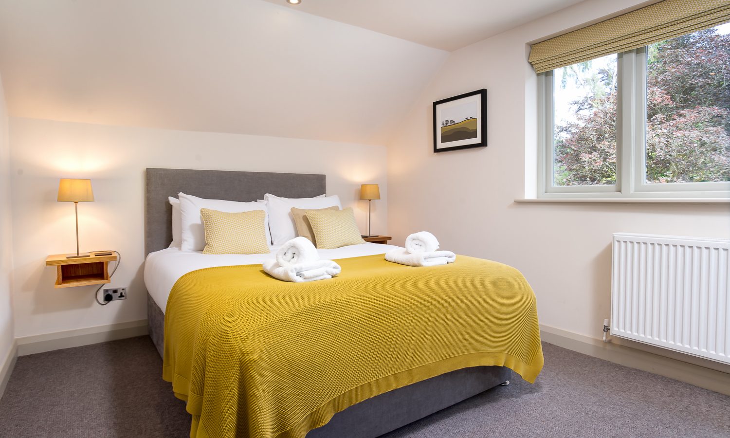 A cozy, well-lit bedroom at Lowfield House features a large double bed adorned with a yellow bedspread and white pillows. Two neatly folded towels are placed on the bed, and natural light streams in from a window flanked by wall-mounted lamps. A framed picture hangs above the radiator.