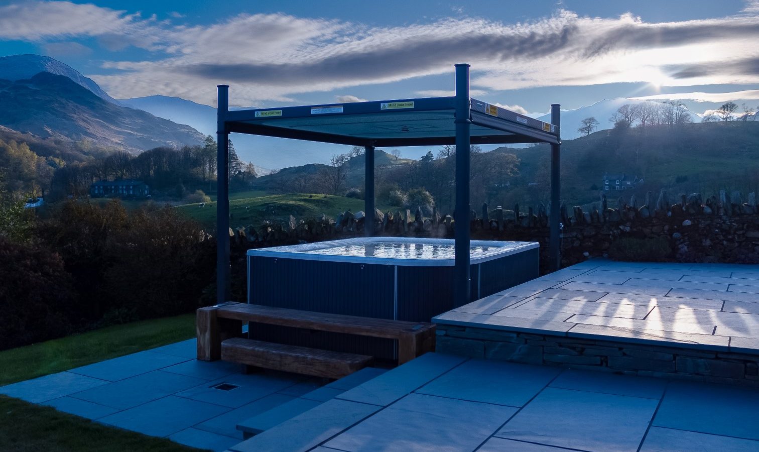 At Lowfield House, a hot tub graces the patio surrounded by stone tiles, overlooking rolling hills and a stone wall. A modern pergola provides coverage, while mountains create an inspiring backdrop under a blue sky with scattered clouds at sunrise or sunset.