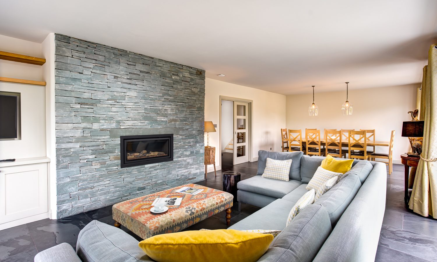 Discover the modern charm of Lowfield House's living room, where a sleek gray sectional sofa adorned with yellow and white pillows invites relaxation. The stone accent wall with a built-in fireplace sets a warm ambiance, while the patterned coffee table on the gray rug completes this stylish space.