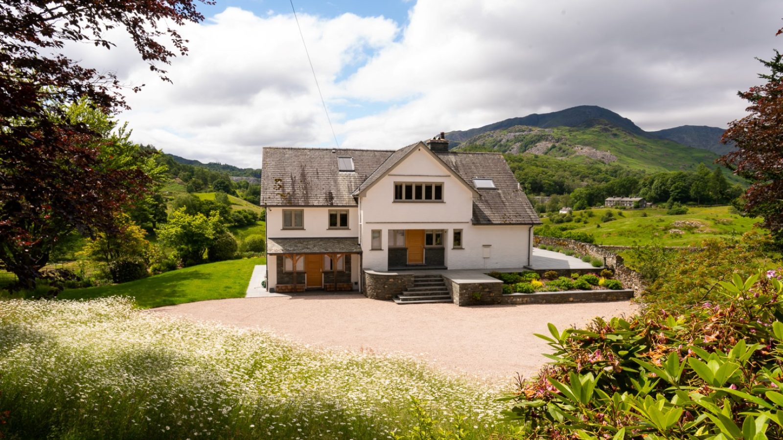 Lowfield House is a large white home with a gray roof, nestled among lush greenery and mountains. A gravel driveway welcomes you, leading to a well-maintained lawn adorned with vibrant flowers. With trees framing the scene under a cloudy sky, this idyllic setting is truly captivating.