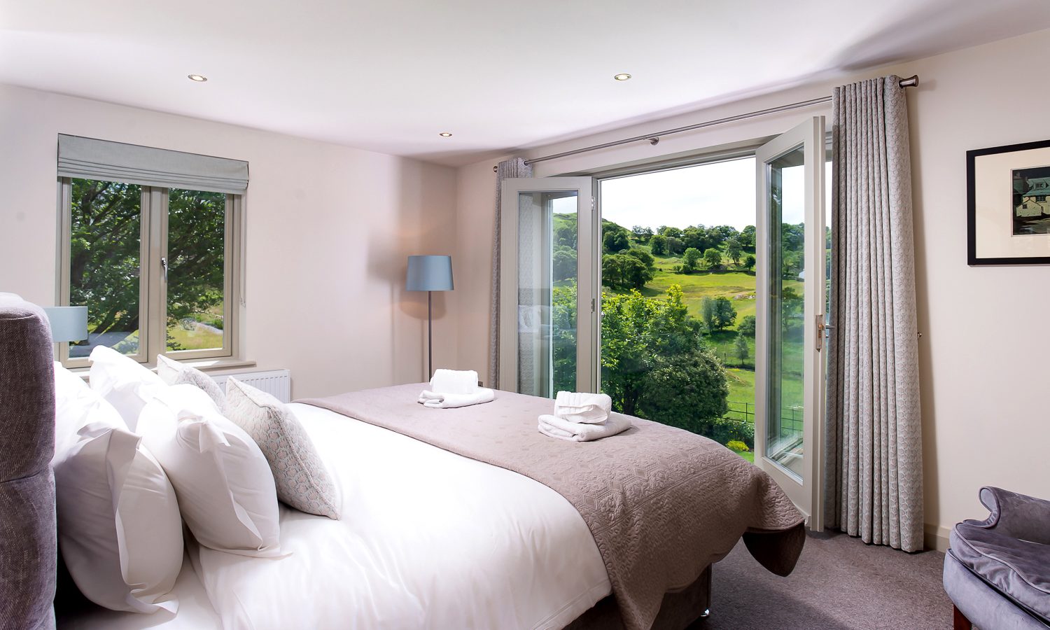 A bright, cozy bedroom at Lowfield House features a large bed with white bedding and soft grey accents. The room has a window and glass doors, opening to a picturesque view of a lush green landscape. A lamp and a framed picture add a touch of elegance to the decor.