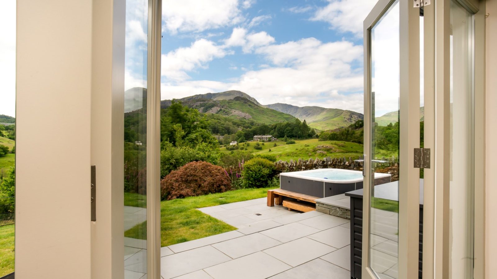 A view from an open doorway at Lowfield House reveals a lush green landscape with hills under a partly cloudy sky. In the foreground, a patio with a hot tub is surrounded by grass and trees, offering a serene escape.