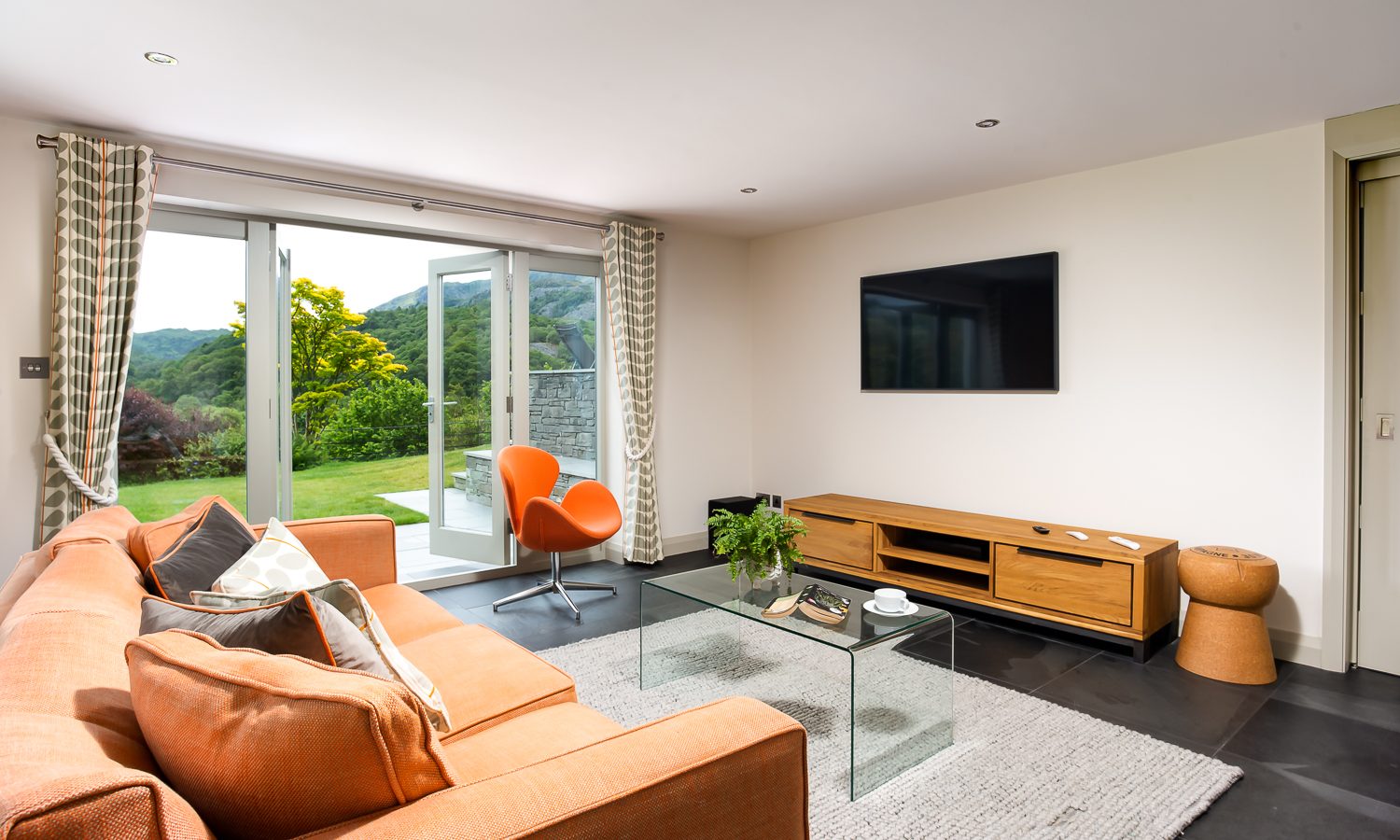 The modern living room at Lowfield House features a large orange sofa, a glass coffee table, and a black flat-screen TV on a wooden stand. A distinctive orange chair sits by sliding glass doors that open to a scenic garden view. The room boasts gray flooring and a light-colored rug.