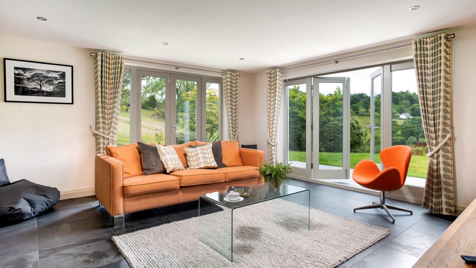 A modern living room in Lowfield House boasts a large glass door opening to a scenic view. It features an orange sofa, a glass coffee table, and an orange chair. The room has a gray rug, slate flooring, and patterned curtains. A framed photo elegantly adorns the wall.