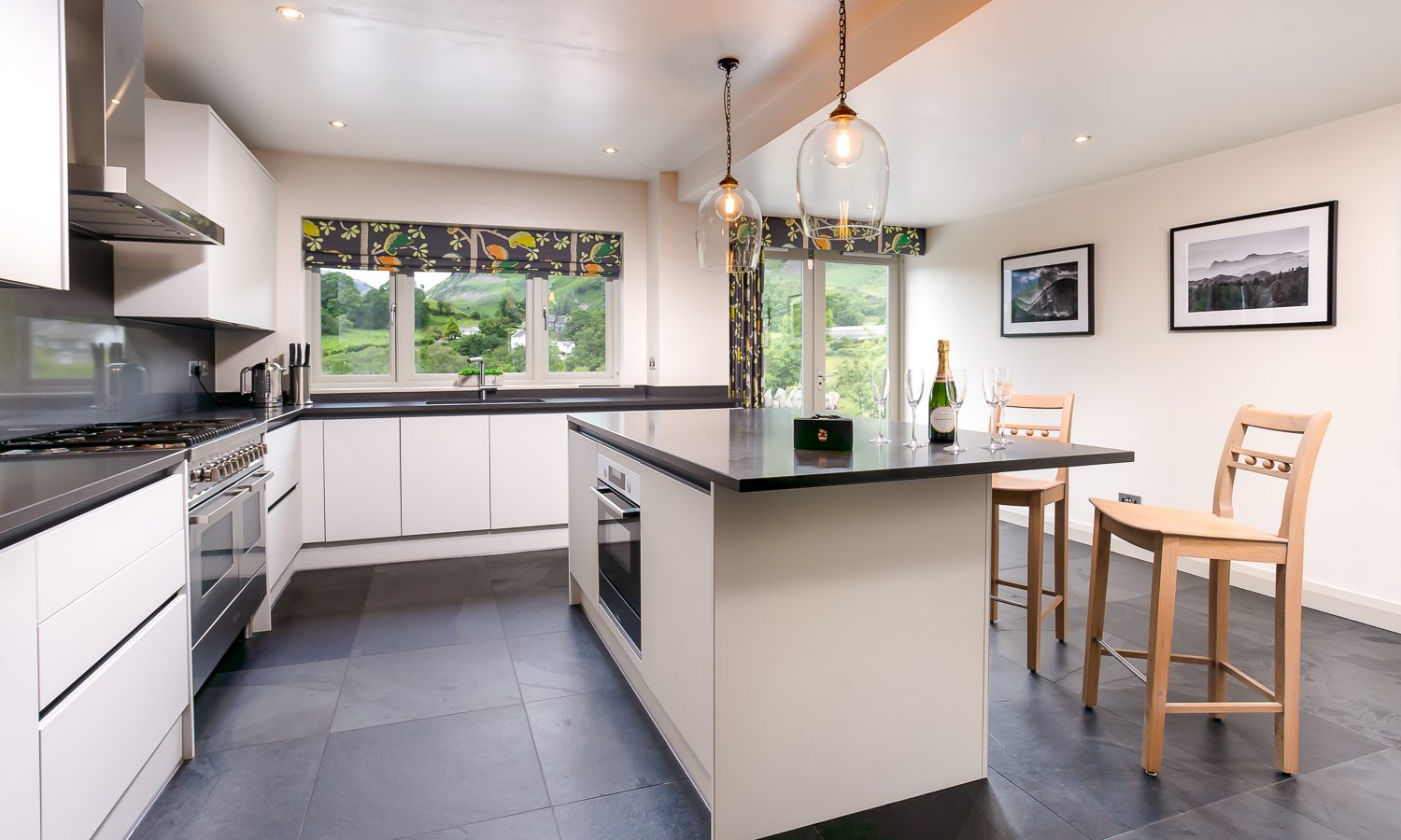 The modern kitchen at Lowfield House features white cabinets, a stainless steel stove, and a central island with two wooden barstools. Pendant lights shine above the island, while large windows offer views of greenery. Decor includes framed pictures and a colorful window valance.