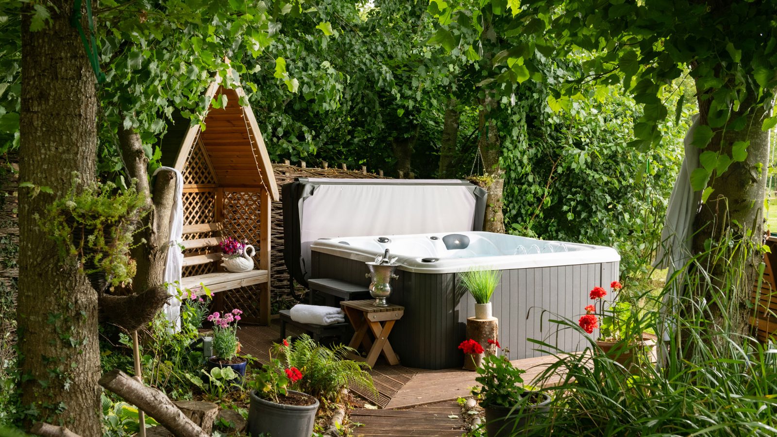 A backyard scene with a grey hot tub surrounded by greenery, evoking the charm of rural retreats. There's a wicker lounge chair with cushions and a side table holding drinks next to the hot tub. Potted plants and flowers add color, and trees provide shade, creating a relaxing atmosphere reminiscent of hop pickers’ havens.