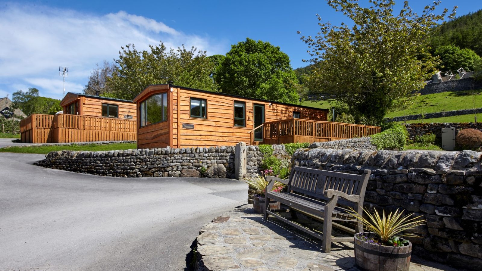 Two wooden cabins sit in a scenic, hilly area with lush greenery and clear blue skies at Howgill Lodge. The cabins are surrounded by stone walls, and a paved path leads to them. A wooden bench with potted plants is in the foreground, creating a peaceful sitting area.