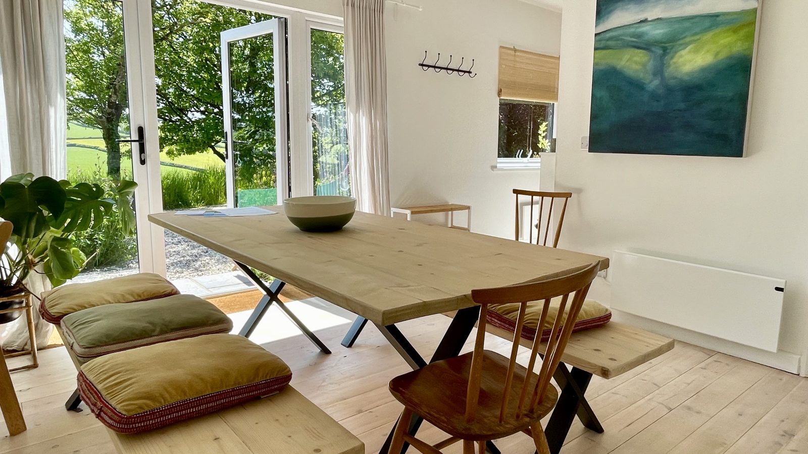 A bright dining room with wooden flooring features a large wooden dining table and matching benches with cushions. Two wooden chairs at the ends of the table add elegance. The room, perfect for holidays, has large glass doors leading outside and a landscape painting depicting the moor on the wall.