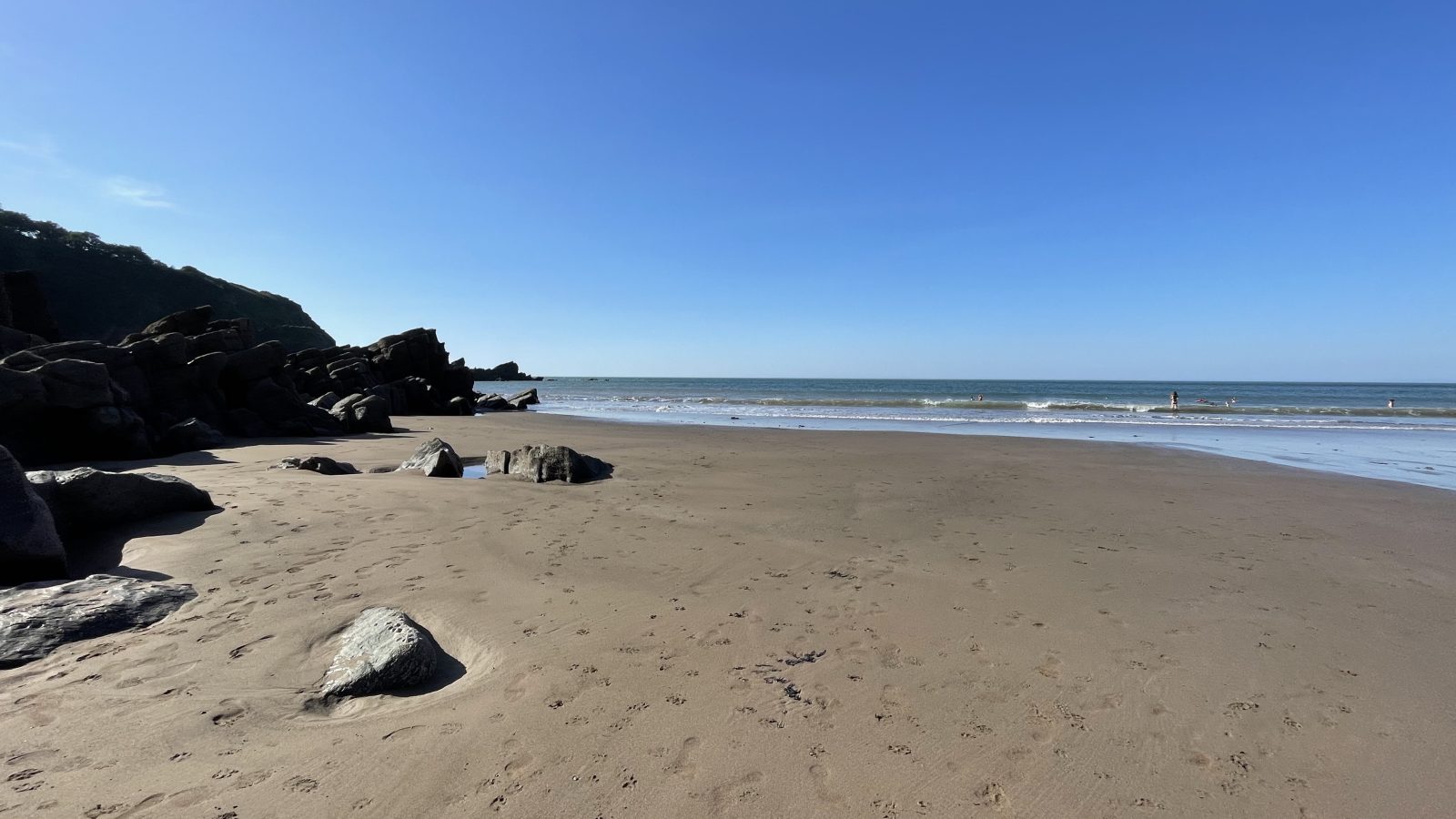 A sandy beach with scattered rocks and a clear sky sets the perfect scene for Moor and Sea Holidays. The ocean waves meet the shore gently, leaving faint human footprints in the sand. On the left, rocky cliffs rise up, partially covered with greenery, while the horizon stretches under a blue sky.