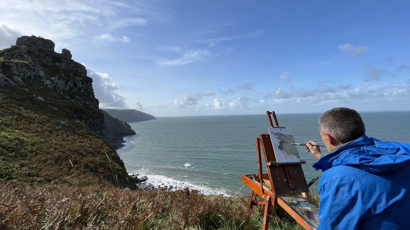 A person wearing a blue jacket is painting on an easel set up on a grassy cliffside. The scene they are painting overlooks a rocky coastline with waves crashing against the shore and a clear blue sky above, capturing the serene beauty of the moor as if on holidays by the sea.