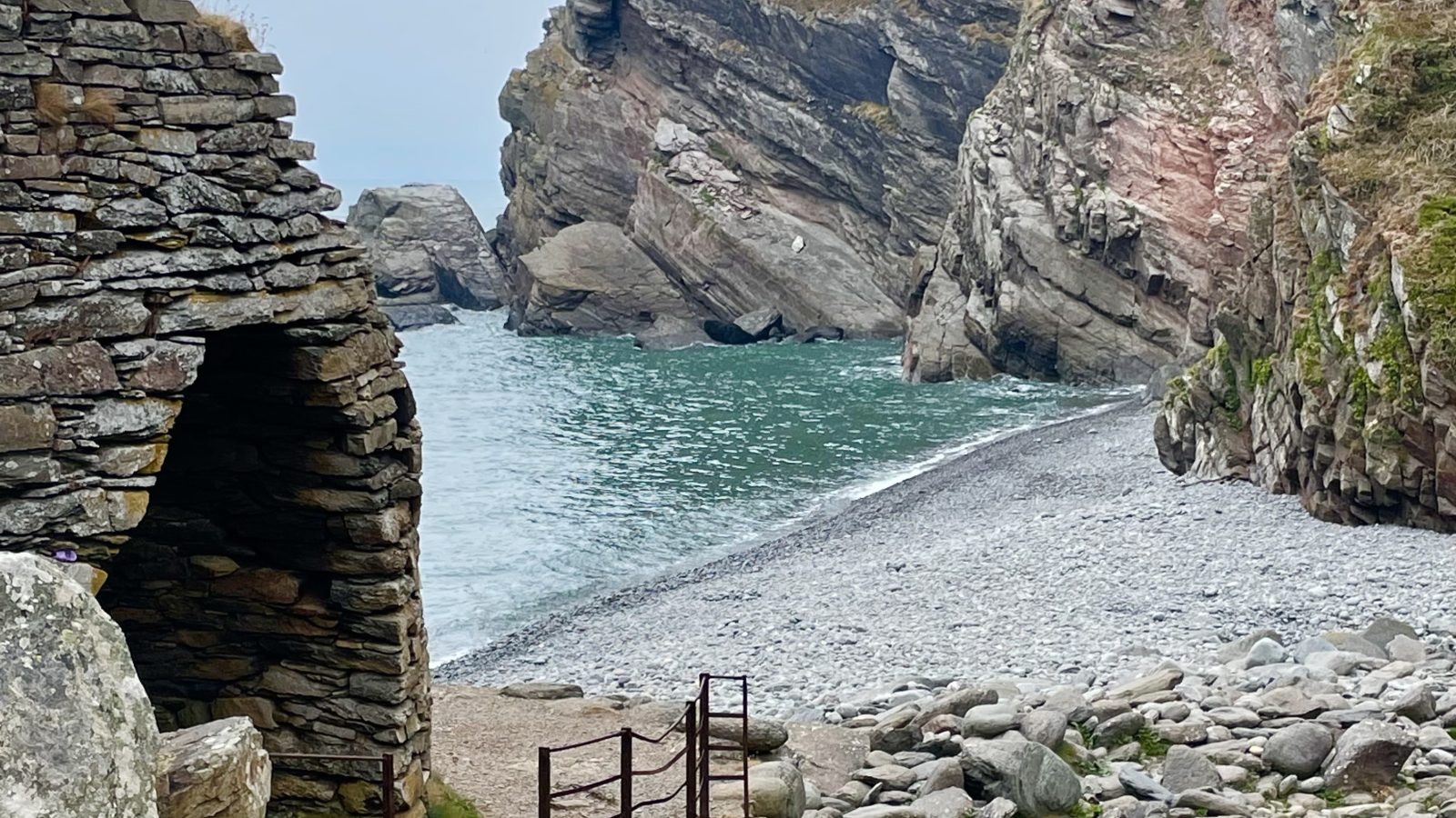 Rocky path leads to a secluded beach with pebbles. On the left, there's an ancient stone structure, and in the background, steep cliffs rise from the sea. The rugged coastline blends gray and pink hues in the rocks under a cloudy sky, perfect for moor-inspired holidays by the sea.