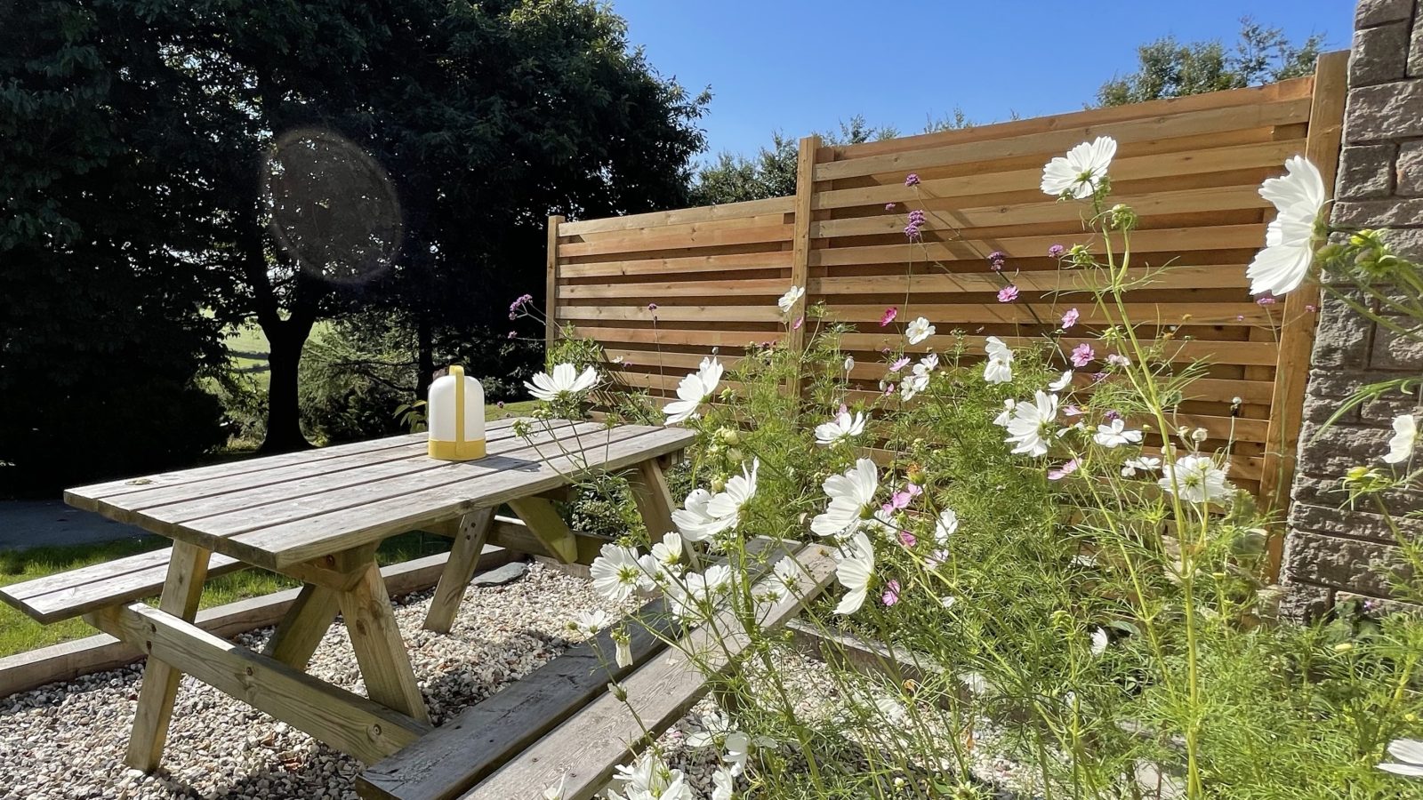 A wooden picnic table with a yellow lantern sits in a sunny garden surrounded by white and pink wildflowers, reminiscent of holidays by the sea. A wooden fence partially encloses the area, with trees visible in the background under a clear blue sky.