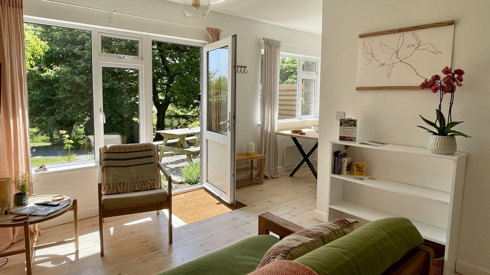 A bright living room with large windows and a glass door opening to a lush outdoor garden. The room features a wooden sofa with green cushions, a wooden chair, a small wooden side table, and a white bookshelf with books and decor inspired by Sea Holidays. A potted orchid adds an elegant touch.